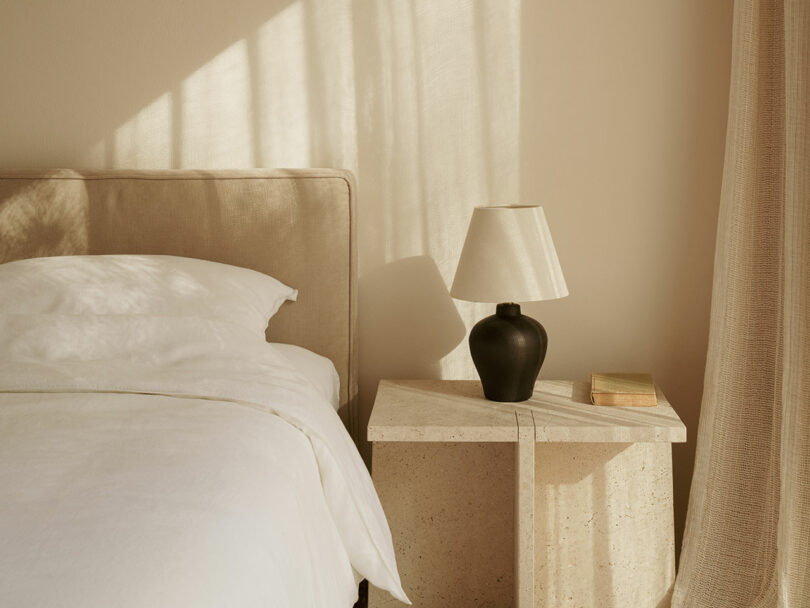 A neatly made bed with white bedding beside a stone nightstand holding a black lamp and a closed book in a softly sunlit beige room.