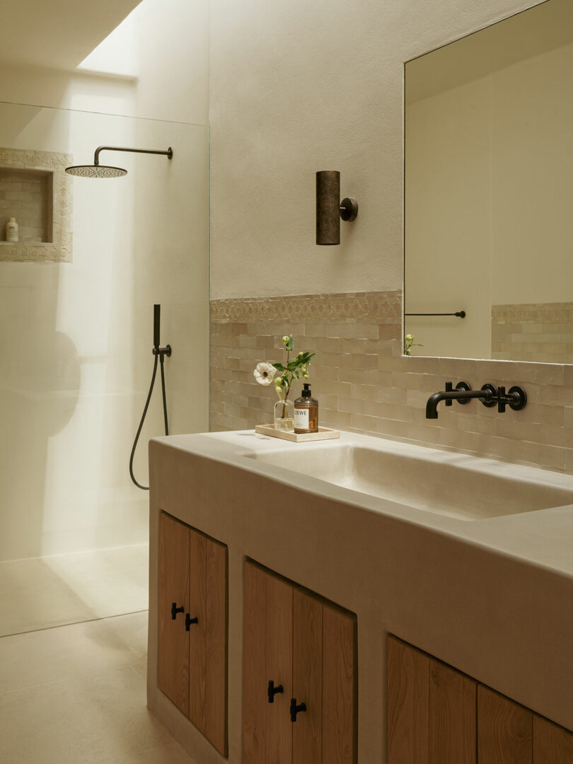 Minimalist bathroom with a walk-in glass shower, wall-mounted black fixtures, a large mirror, wooden cabinet doors, and a small vase with flowers on the countertop.