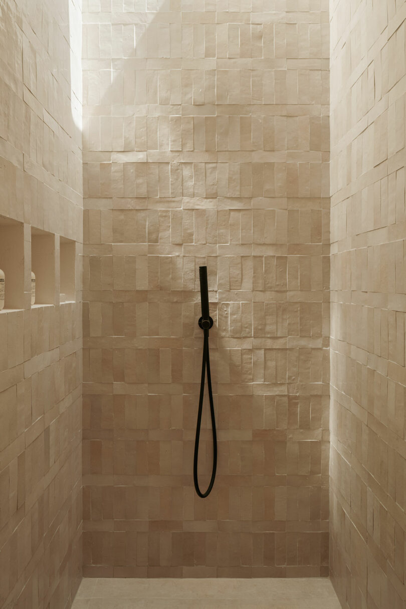 Minimalist shower with textured beige tile walls, a black handheld showerhead, and two built-in shelf niches containing bottles.