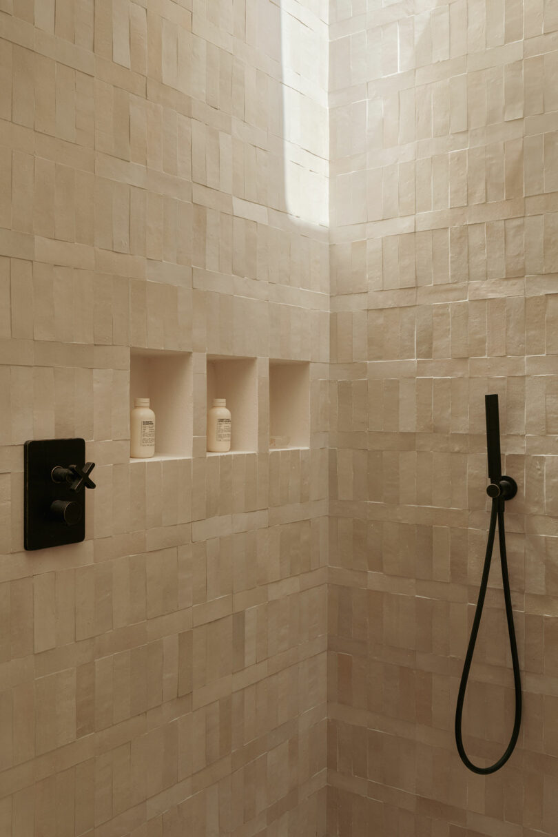 A tiled shower with three built-in shelves holding bottles, a black wall-mounted control panel, and a black handheld showerhead.