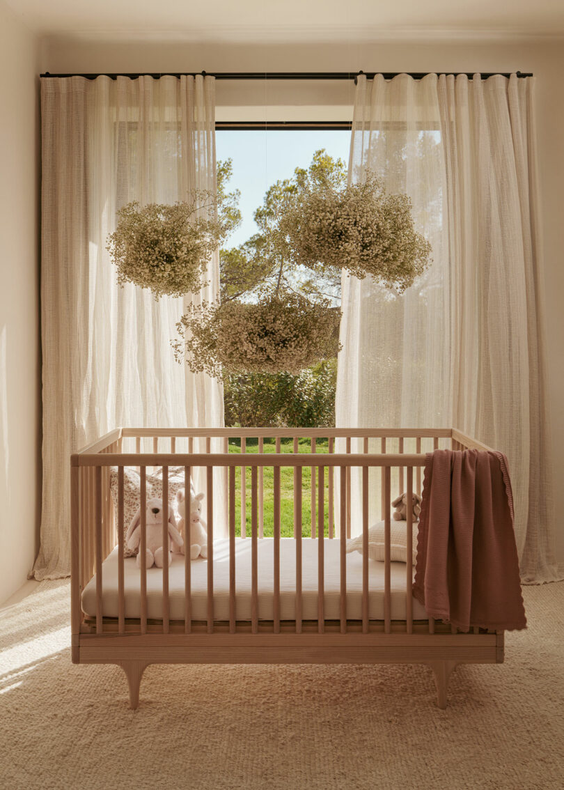 A wooden crib with stuffed animals and a folded blanket stands in front of sheer curtains and a large window with hanging floral arrangements and trees visible outside.
