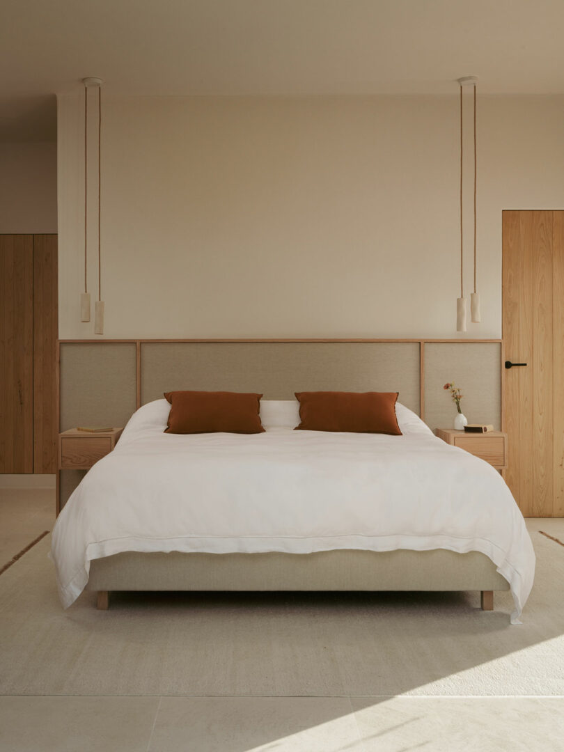 Minimalist bedroom with a double bed, white bedding, two brown pillows, pendant lights, wooden nightstands, and light wood doors and panels.
