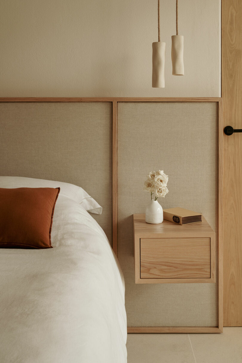 Minimalist bedroom with a beige and wood nightstand, white bedding, a rust-colored pillow, a small vase of white flowers, a closed book, and two pendant lights.