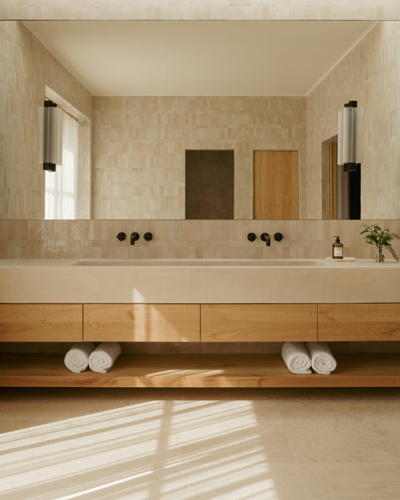 Modern bathroom with light tile walls, large mirror, double sinks with black faucets, wooden vanity, rolled white towels, and natural light from a window.