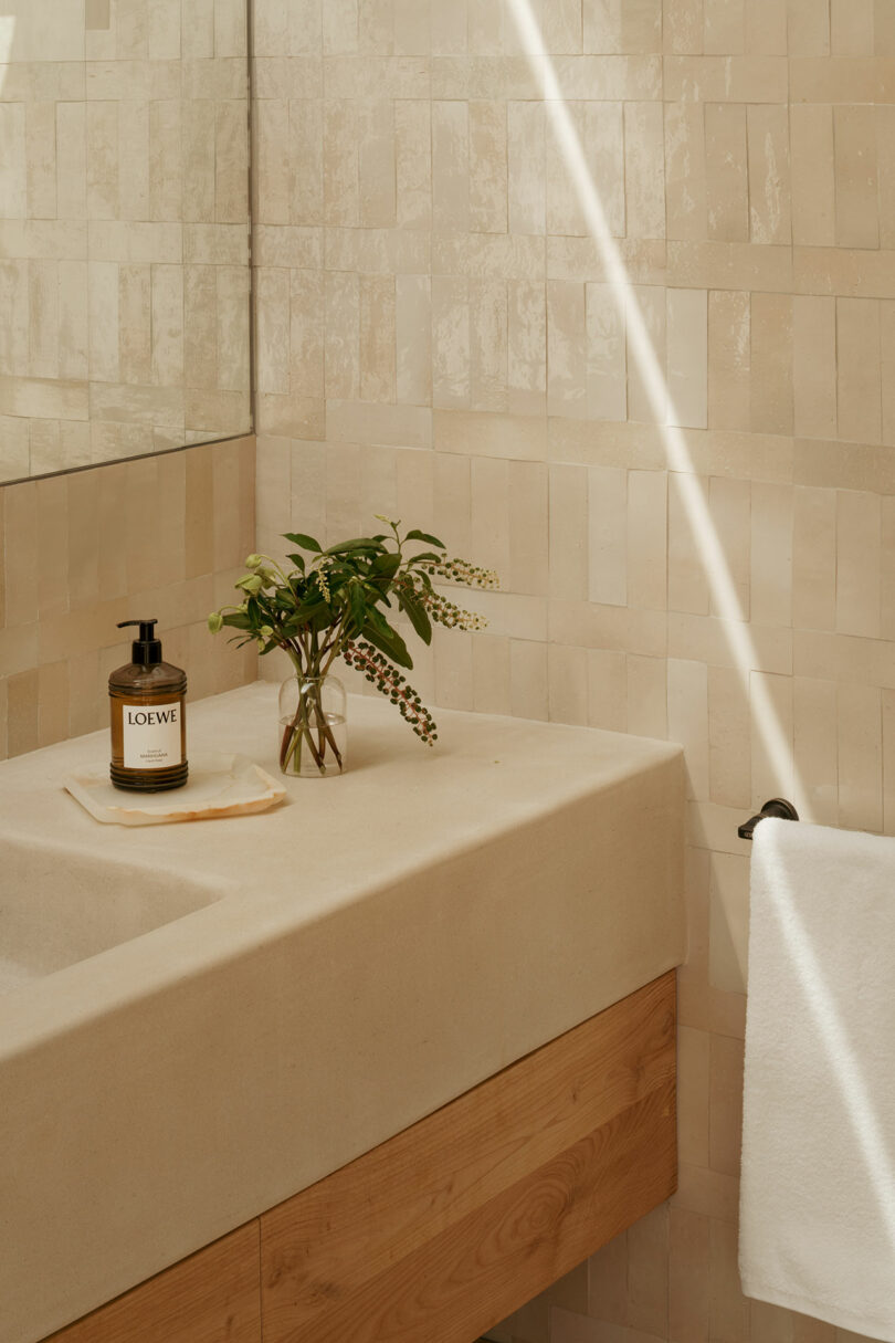 A beige tiled bathroom features a stone sink with a wooden base, a bottle of LOEWE hand soap, a small vase with greenery, and a white towel on a rack. Sunlight streams in.