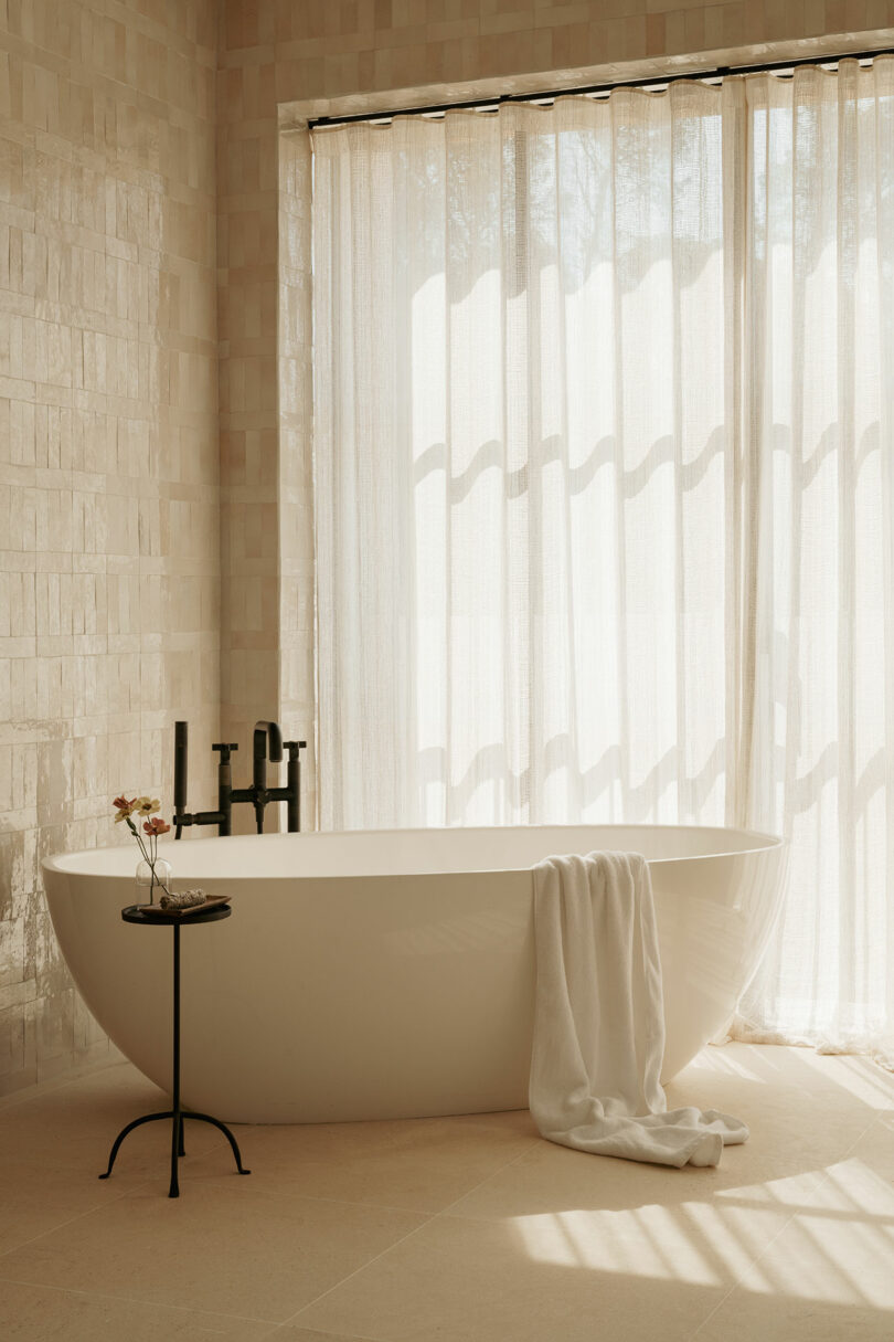 A modern bathroom with a freestanding white bathtub, a towel draped over the edge, a small round side table, and sheer curtains letting in natural light.