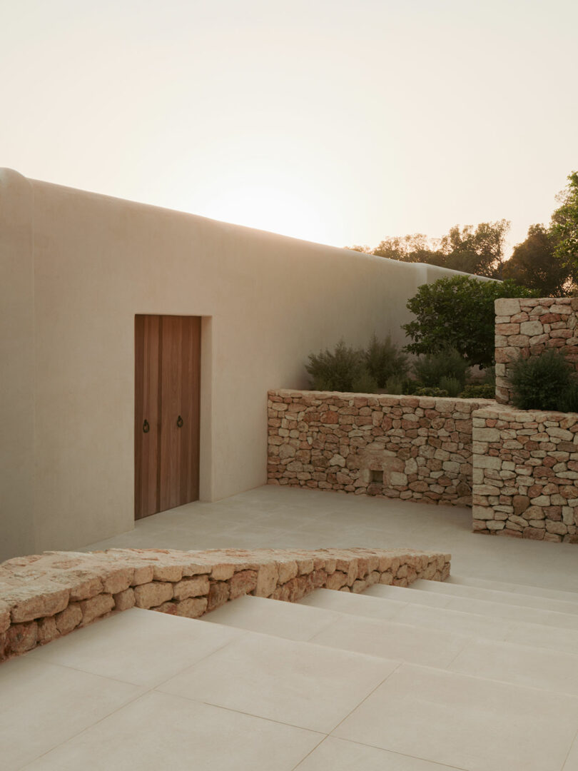 Minimalist stone staircase and walls with a wooden door, surrounded by greenery, under warm evening sunlight.