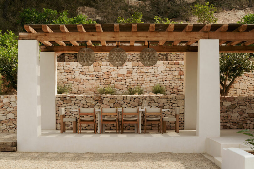 Outdoor dining area under a wooden pergola with a long table, chairs, hanging lights, and a stone wall backdrop.