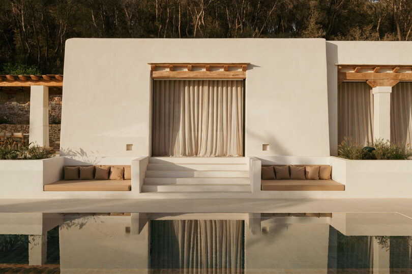 Modern white stucco building with beige curtains, wooden accents, built-in seating, and steps leading to a pool reflecting the structure.