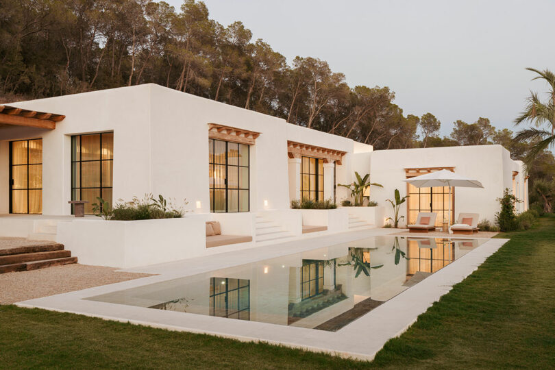Modern white villa with large windows, a rectangular swimming pool, poolside loungers, and surrounding greenery, set against a backdrop of trees.