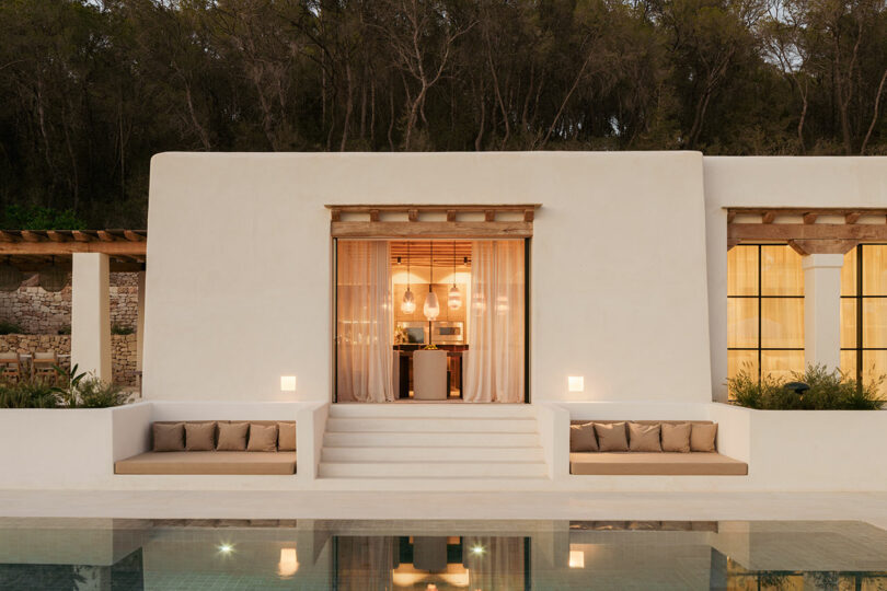 Modern villa exterior with white stucco walls, symmetrical built-in seating, large windows, central steps, and a reflection in a pool, set against a backdrop of trees.