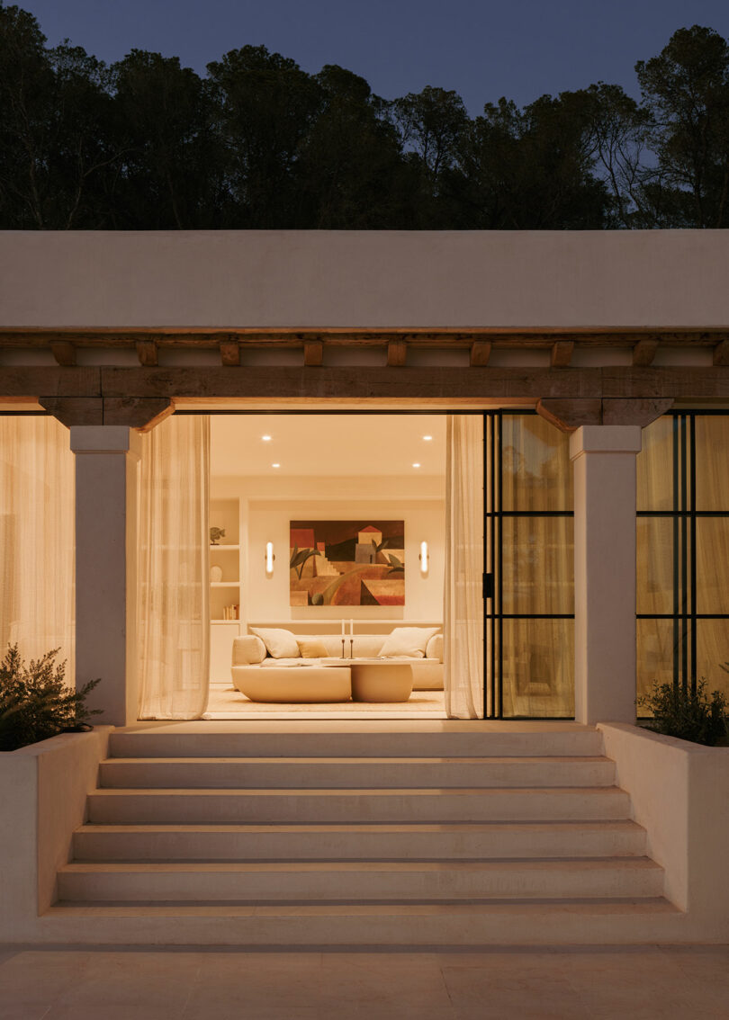 A modern, warmly lit living room is visible through large glass doors, with light-colored sofas, a geometric painting, and steps leading up to the entrance at dusk.