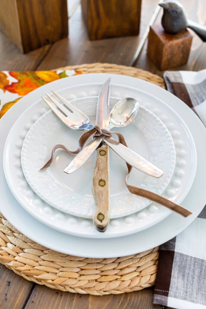 Table place setting of a white dinner plate layered with a fork, knife and spoon tied with brown ribbon.