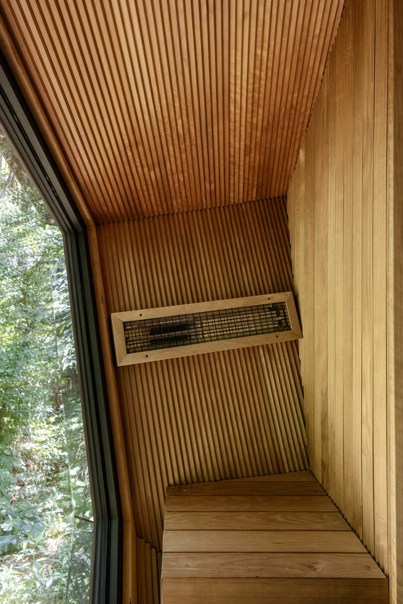 Wood-paneled interior wall and ceiling with a heater vent, next to a large window overlooking green trees outside.