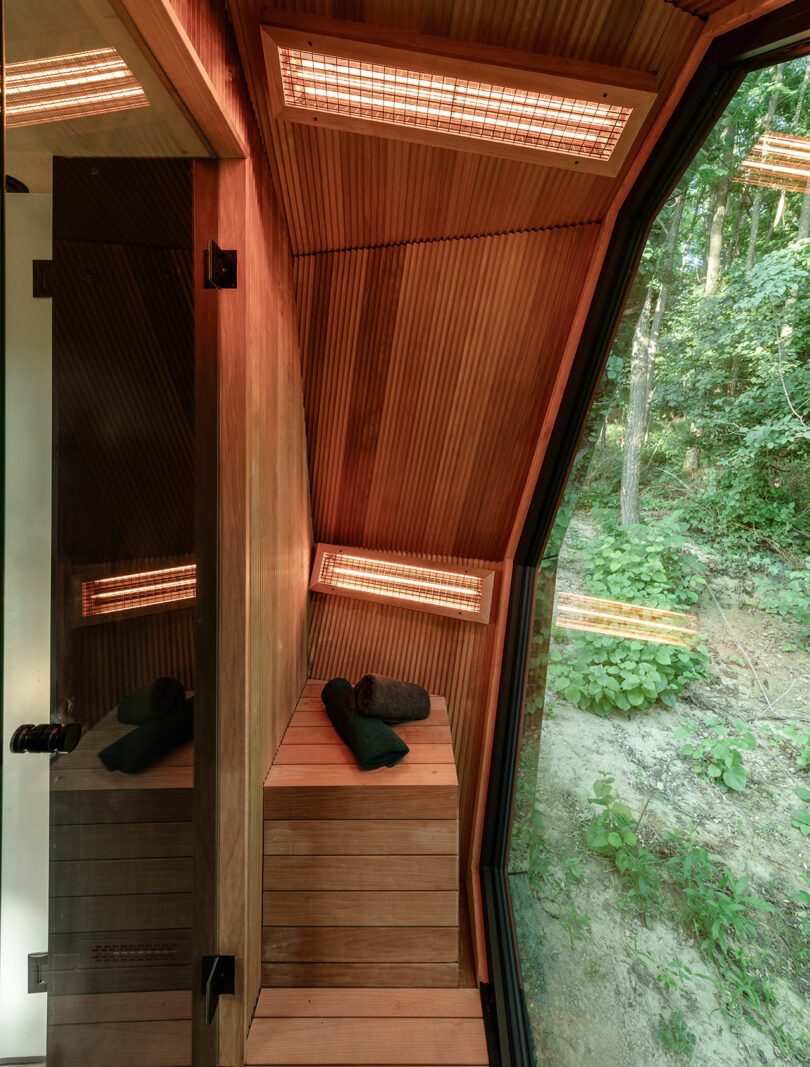 A modern wooden sauna with built-in heaters, a bench, and two rolled towels, featuring a large window overlooking a forested area.