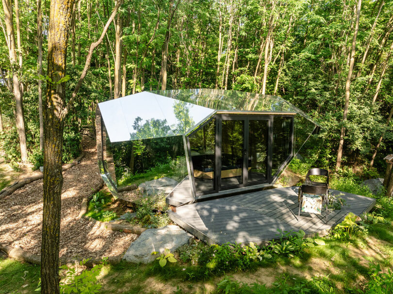 Modern cabin with mirrored exterior walls and large glass doors, situated on a wooden deck in a lush, green forest clearing.