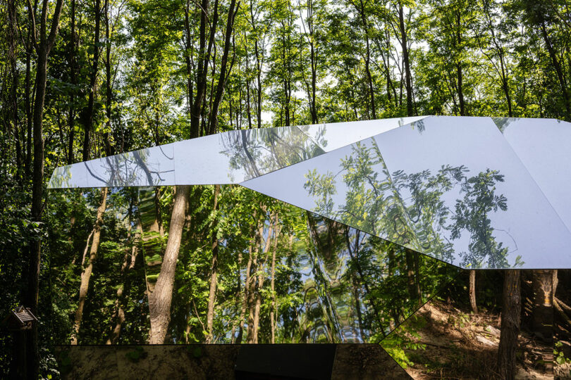 A mirrored building reflects trees and greenery in a dense forest, blending into its surroundings with angular, geometric surfaces.