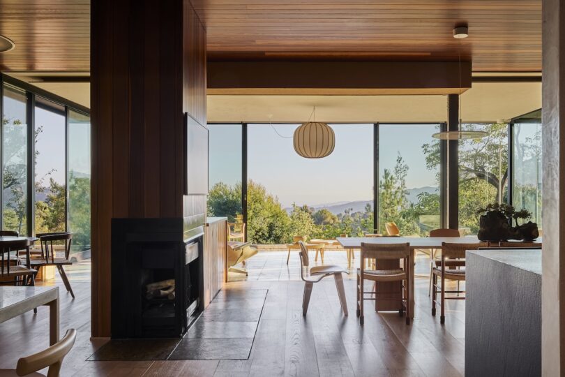 Modern open-plan living and dining area with wooden furnishings, floor-to-ceiling windows, and a view of trees and distant hills under clear skies.