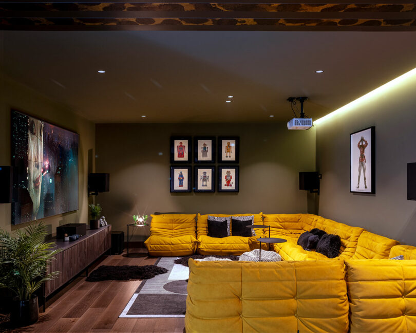 A modern home theater room with a large yellow sectional sofa, wall-mounted TV, framed artwork, projector, and indoor plants on wooden flooring.