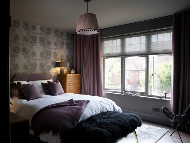 A neatly made bed with purple pillows and a blanket, a wooden dresser, large window with blinds, and patterned wallpaper in a softly lit bedroom.