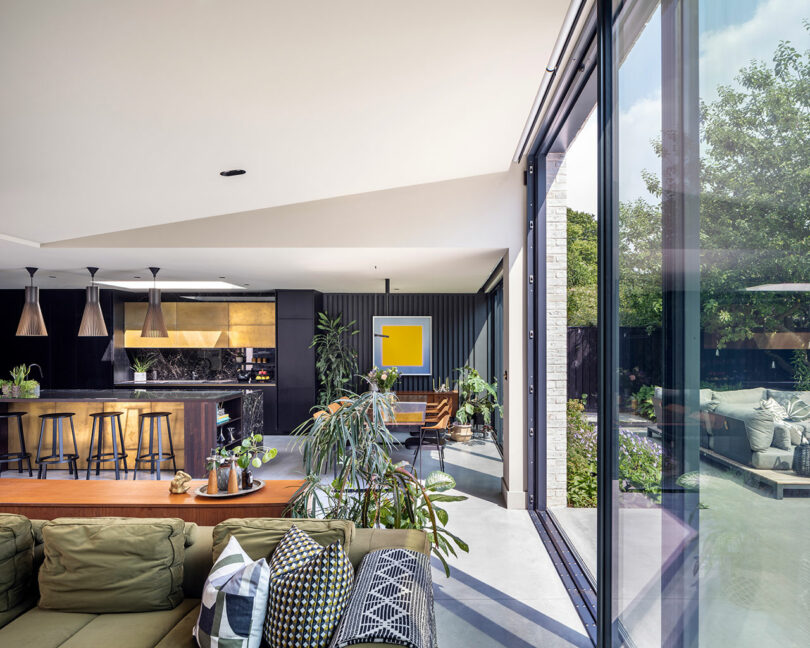 Modern open-plan living area with green sofa, kitchen island, indoor plants, and large glass doors opening to a patio with outdoor seating and garden.