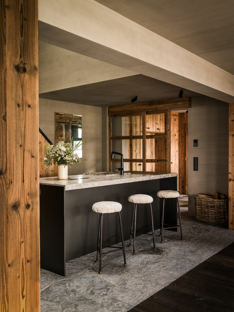 Modern kitchen with a black island, three cushioned bar stools, stone flooring, wooden walls, open shelves, and a vase of flowers on the counter.