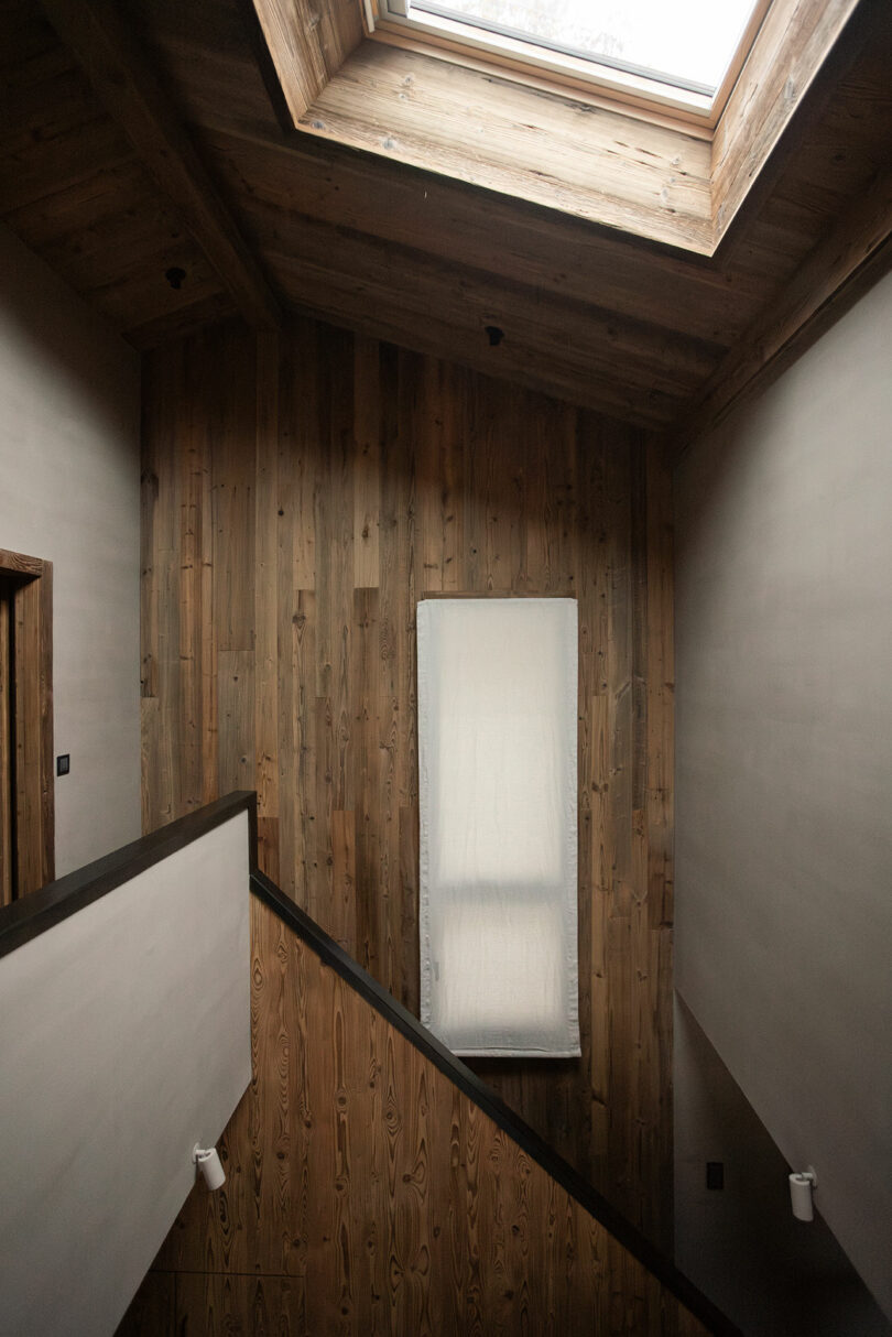 A wooden staircase landing with a large skylight above and a narrow covered window on the wall, featuring natural wood paneling and minimal decor.