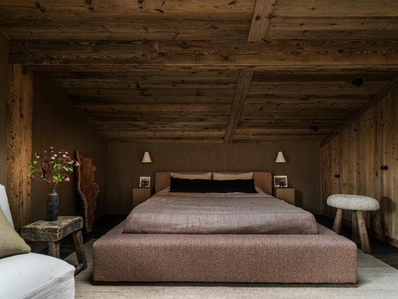 A cozy bedroom with a large bed, wooden ceiling and walls, two bedside tables with lamps, a stool, and a vase of flowers on a rustic side table.