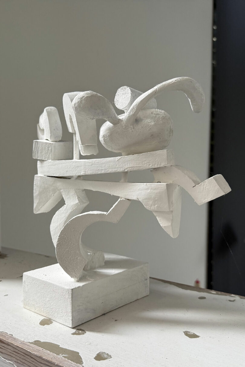 Abstract white sculpture with intersecting geometric and curved shapes, displayed on a white base against a plain background.