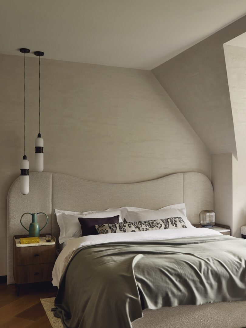A modern bedroom with a curved upholstered headboard, two pendant lights, a neatly made bed with gray bedding, and wooden nightstands with decorative items.
