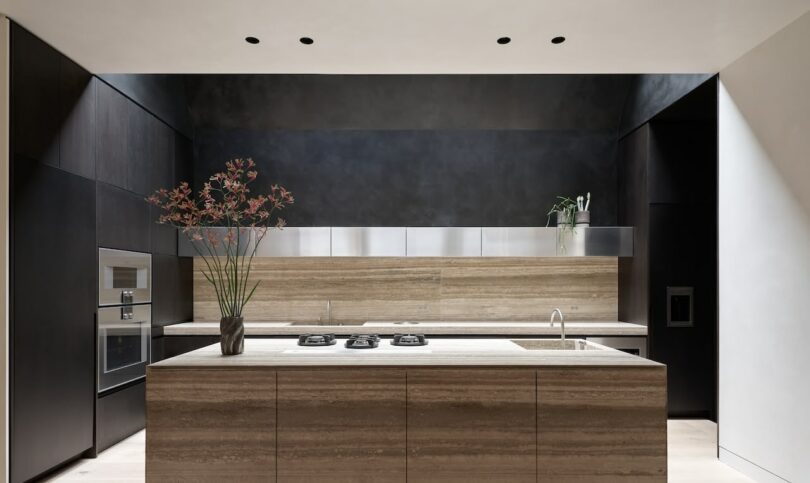 Modern kitchen with a central island, built-in appliances, dark cabinetry, metal backsplash, and decorative plants on the counter—perfect for enjoying a morning coffee or a glass of skim milk in style.