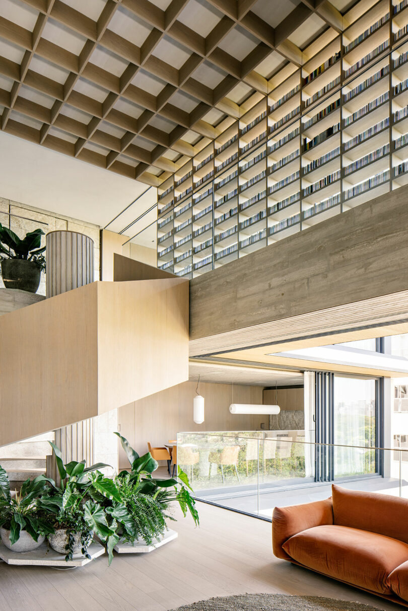 Modern interior with a geometric ceiling, large bookshelf, open staircase, indoor plants, and orange sofa in a bright, spacious living area with floor-to-ceiling windows.