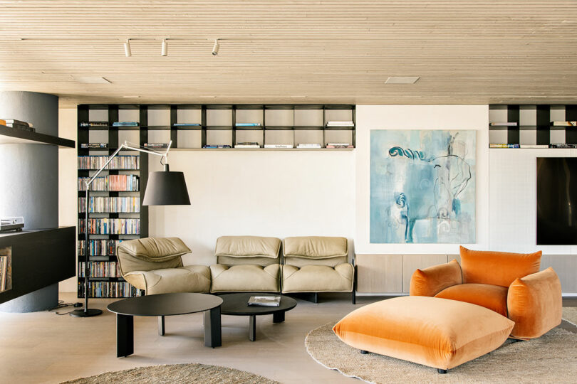 A modern living room with beige lounge chairs, an orange armchair with ottoman, black shelves, a floor lamp, a bookshelf, and a large abstract blue painting on the wall.