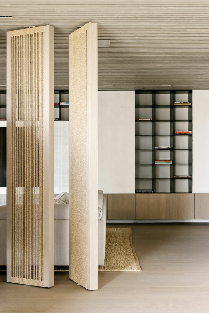 Minimalist living room with light wood floors, a beige woven rug, two vertical room dividers, a grey sofa, and built-in shelving along the back wall with a few books.