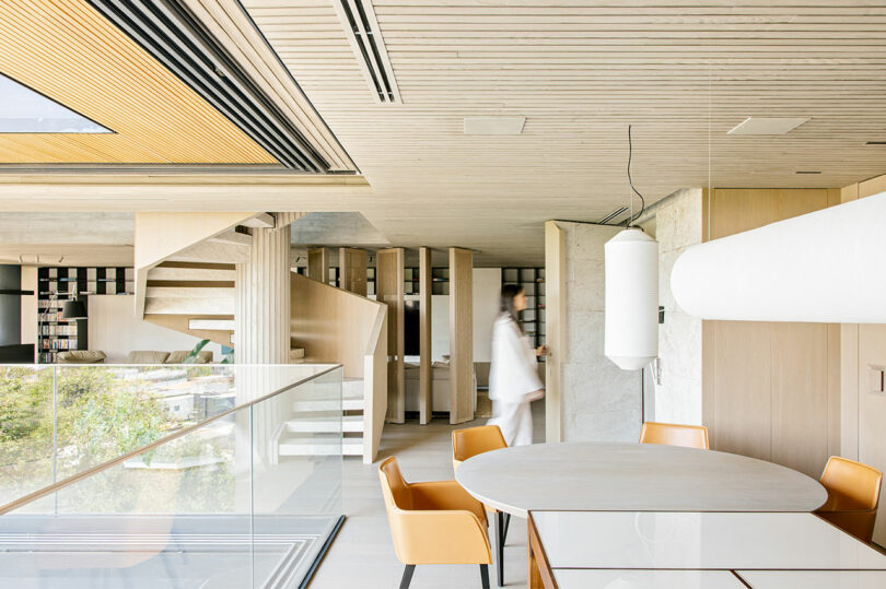 Modern dining area with round table and four chairs, glass railing, and a blurred person walking in a bright, minimalist interior with wooden finishes and staircase in the background.