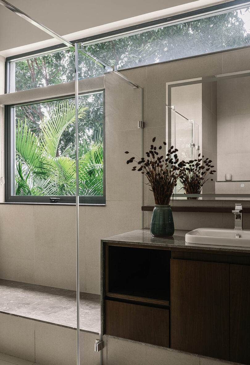 Modern bathroom with a large window showing green foliage, a countertop with a white sink, dark wood cabinet, and a vase of dried plants.