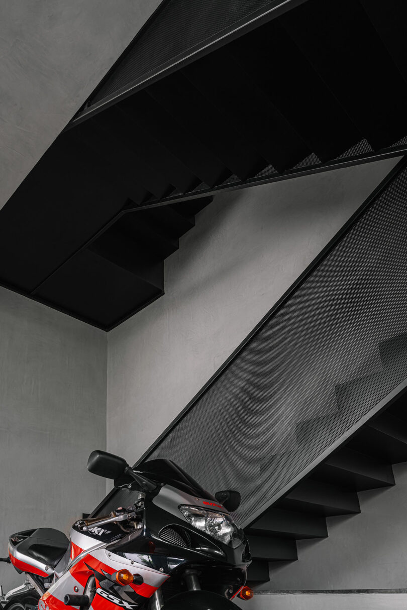 A black and red motorcycle is parked indoors next to a modern, black metal staircase with geometric lines against a gray wall.