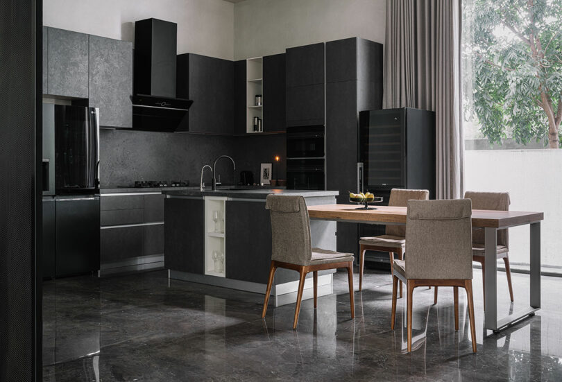 Modern kitchen with dark cabinets, built-in appliances, a dining table with four chairs, and large windows with drapes overlooking a tree outside.