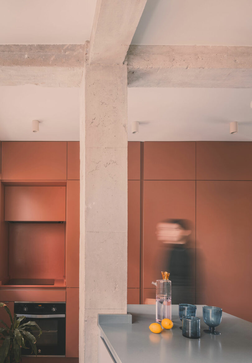 A modern kitchen with matte terracotta cabinets, a concrete pillar, glassware, and lemons on a countertop; a blurred person stands in the background.