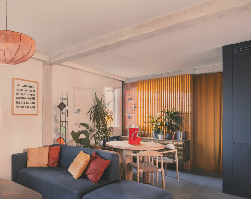 Modern living and dining area with a blue sectional sofa, colorful cushions, round dining table, indoor plants, and large yellow curtains.