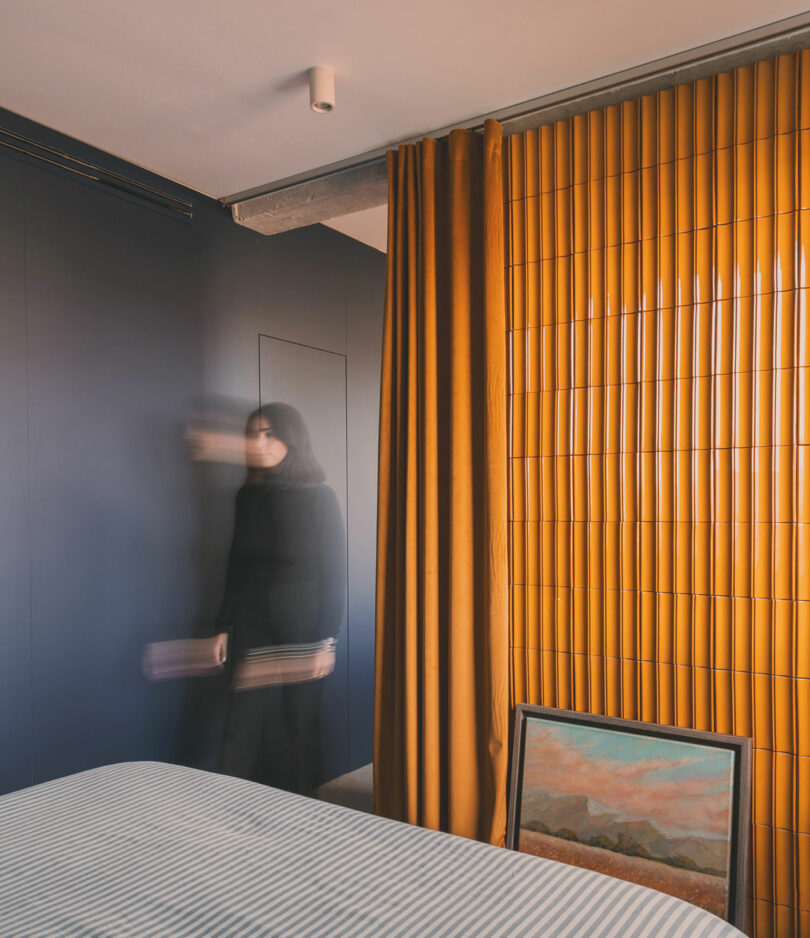 A blurred person in black stands near a dark wall in a bedroom with yellow vertical blinds, a curtain, and a framed painting resting on the floor.