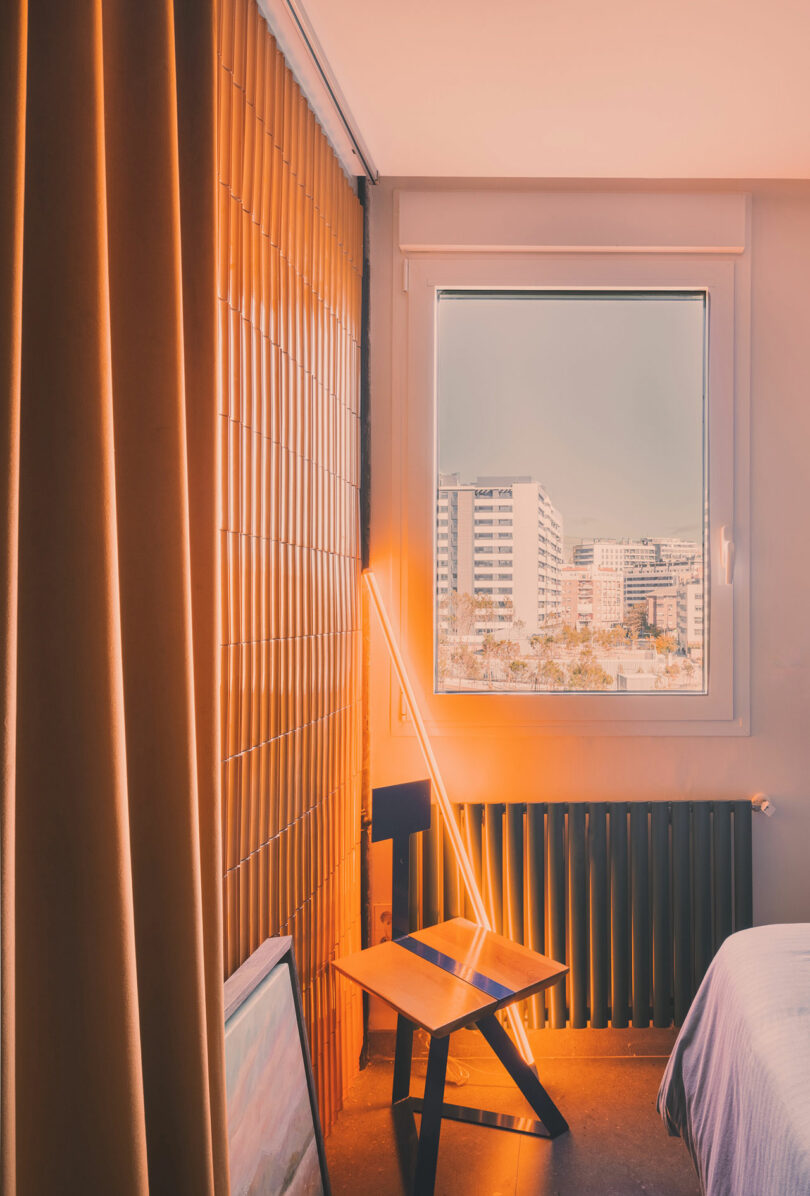 A modern bedroom corner with a geometric chair, a floor lamp, a window showing city buildings, and orange-toned lighting.