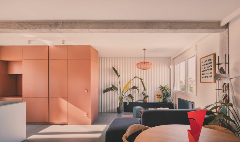 Modern living space with peach-colored cabinetry, gray seating, large indoor plants, and ample natural light from wide windows.