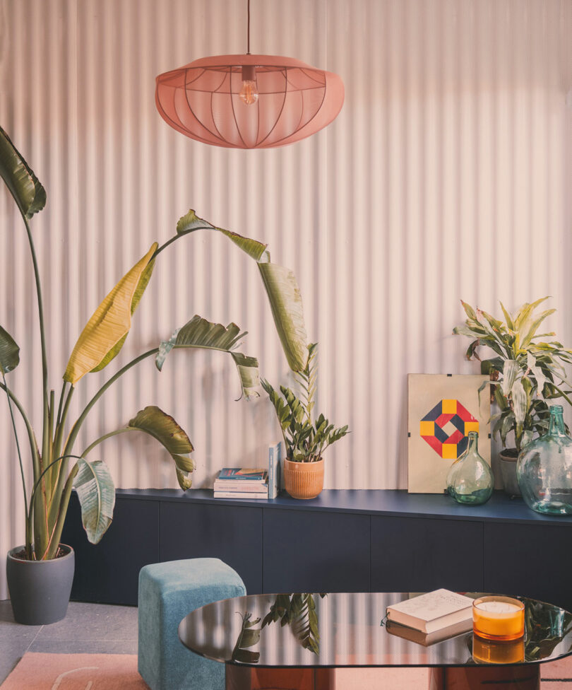 Modern living room with large potted plants, books, a geometric artwork, glass vases, a pink pendant light, and a reflective coffee table with a drink and books.