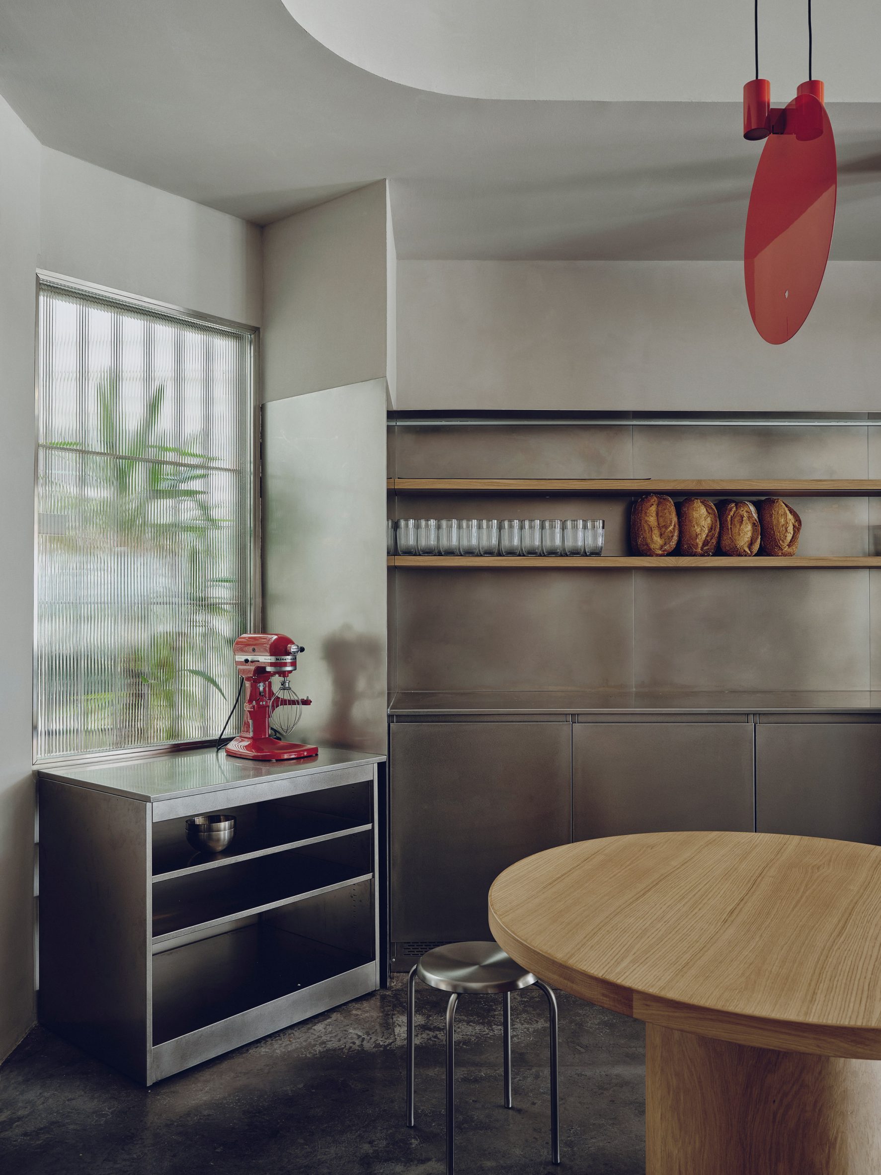Minimalist stainless-steel kitchen with red stand mixer, ribbed glass window, wooden shelves holding glasses and bread, and round wooden table.