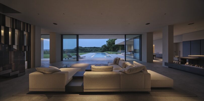 Modern living room with a sectional sofa facing large glass doors that open to a pool and patio, with a scenic outdoor view at dusk.