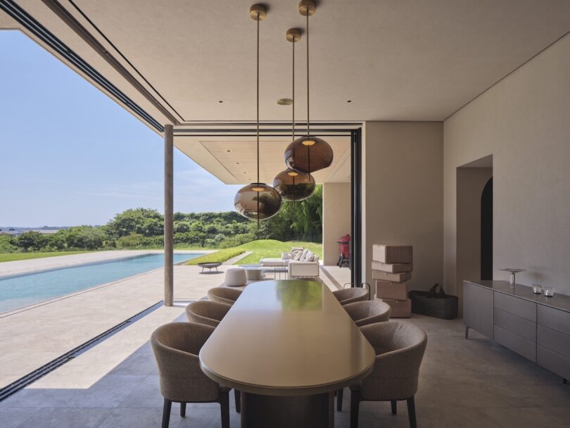 Modern dining area with a long table and chairs, pendant lights above, and large open doors leading to a pool and garden view; several boxes are stacked near the wall.