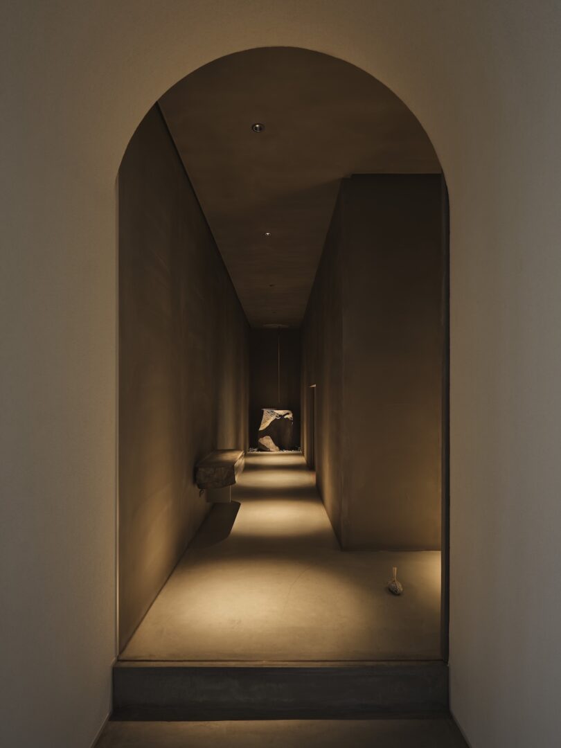 A dimly lit, narrow hallway with smooth brown walls, arched entryway, and a single stone bench along one side, leading to a softly illuminated area at the end.
