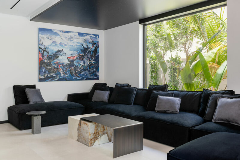 Modern living room with a large black sectional sofa, marble and metal coffee table, abstract wall art, and a large window showing green plants outside.