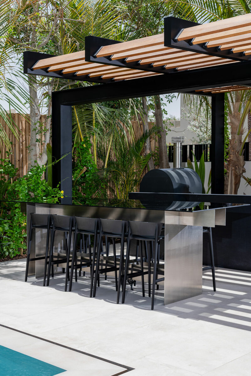 Outdoor kitchen with a modern stainless steel bar, six black bar stools, a grill, and a pergola, set beside a pool and surrounded by lush greenery.